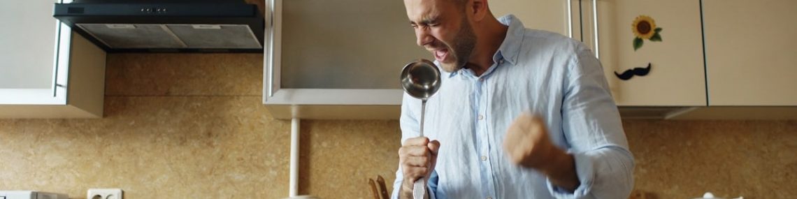 Attractive young funny man dancing and singing with ladle while cooking breakfast in the kitchen at home in the morining